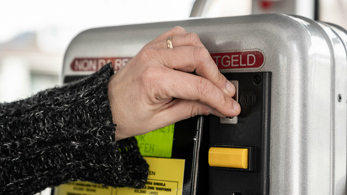 A person is buying a ticket at a ticket machine on the bus. You can see a hand, which is inserting a coin in the machine.