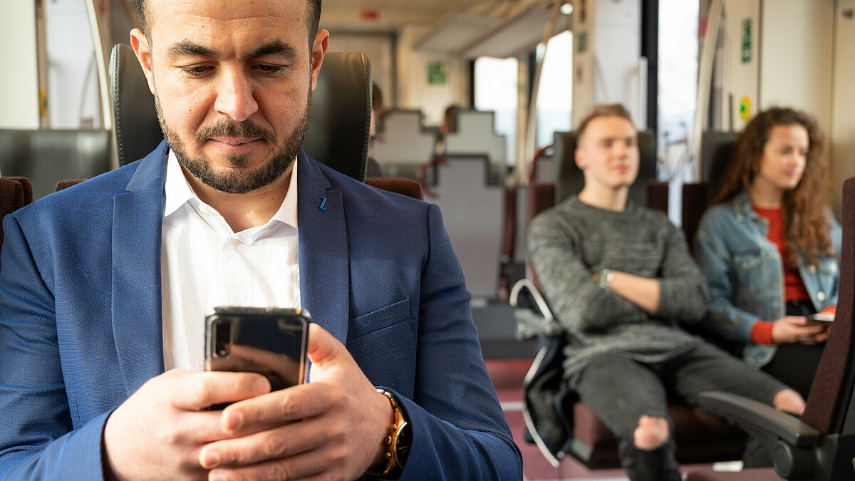 A passenger on the train is checking his phone. You can see a couple on the background.